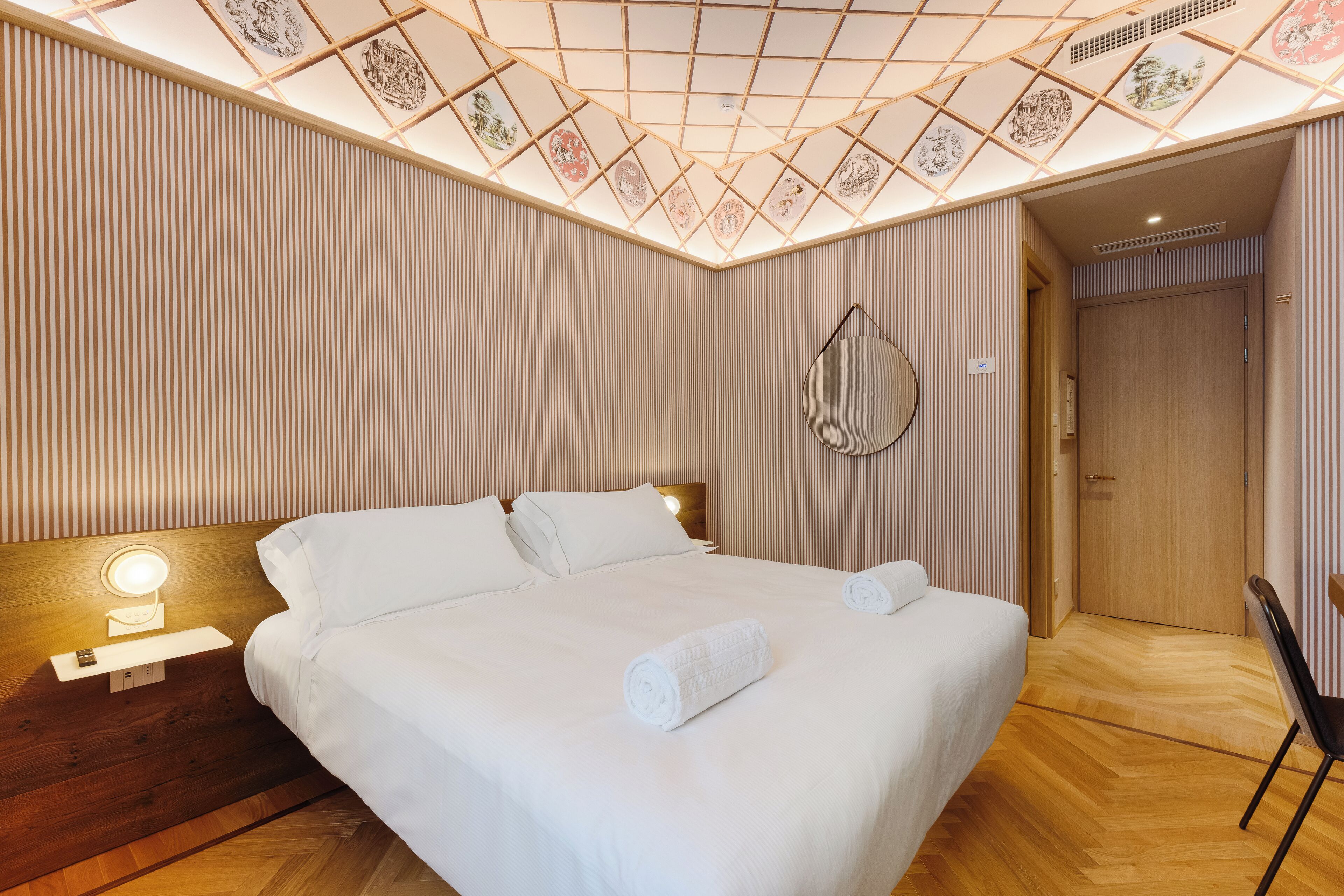 Une chambre d’hôtel avec un grand lit, une table de chevet avec une lampe, une tête de lit en bois, un miroir fixé au mur et une porte donnant sur une autre pièce.