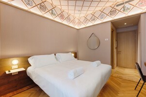 Een hotelkamer met een groot bed, een nachtkastje met een lamp, een houten hoofdeinde, een spiegel aan de muur en een deur naar een andere kamer.