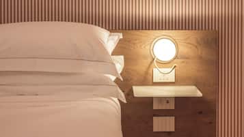 Una cama bien hecha con sábanas blancas, una mesita de noche con un espejo redondo y un enchufe en la pared.