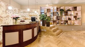 Zona de recepción con un mostrador de madera, un espejo y un papel tapiz floral. Se ven estantes con varios objetos y una planta en maceta.