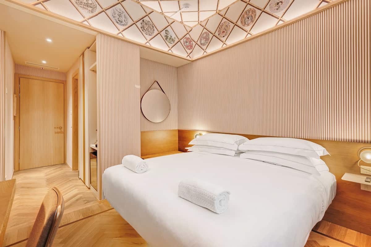 Une chambre d’hôtel avec un grand lit, un parquet en bois, un plafond orné d’un motif géométrique et une armoire en bois.