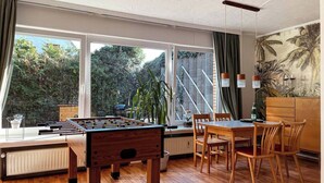 Une pièce équipée d’un baby-foot, d’une table à manger, de chaises et offrant une vue sur la verdure grâce à de grandes fenêtres.
