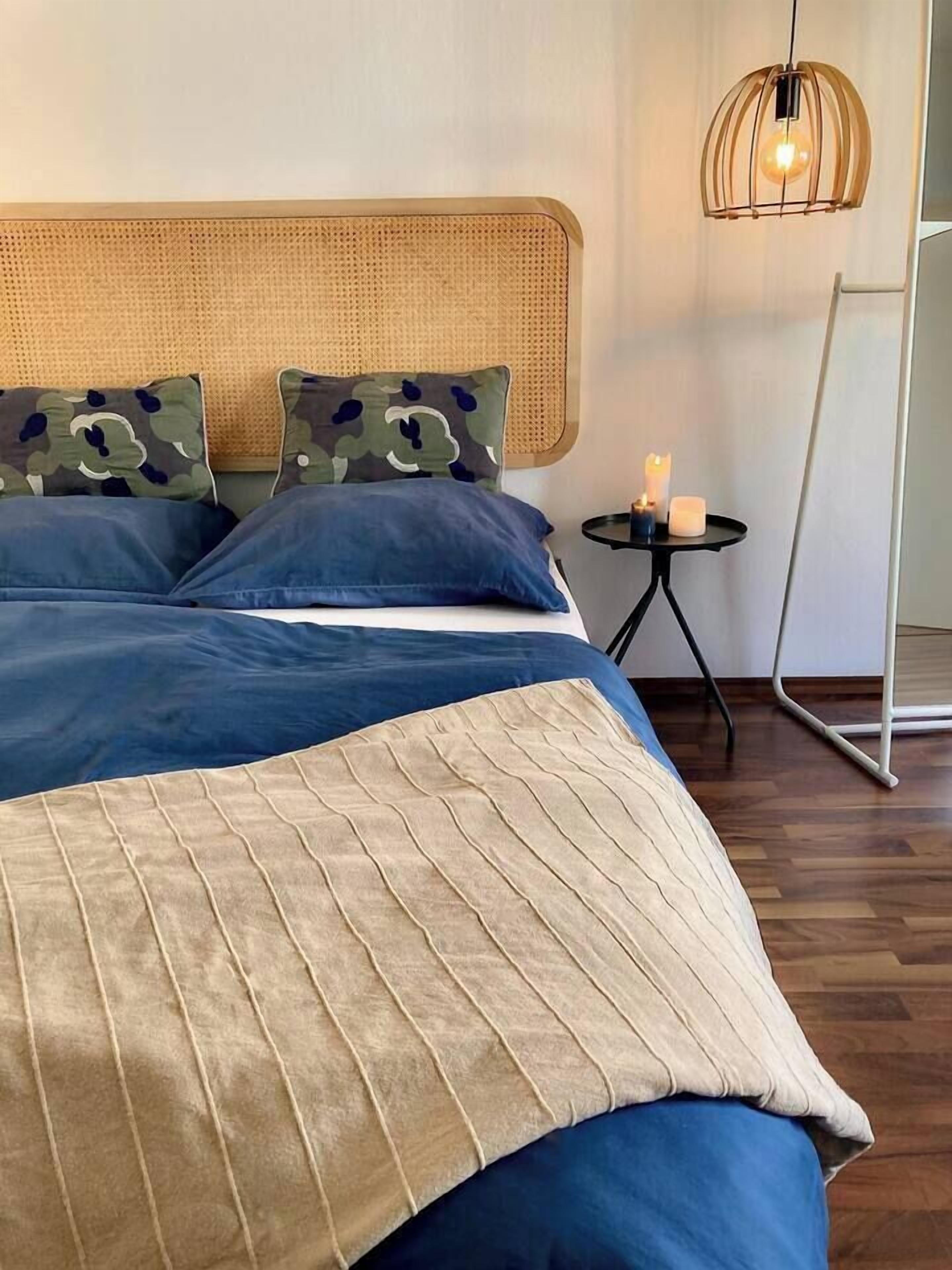 Ein ordentlich bezogenes Bett mit blau-beiger Bettwäsche, einem Rattan-Kopfschild und einem kleinen Nachttisch mit Kerzen.