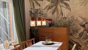 Un coin repas avec une table en bois, des chaises, un petit espace cuisine avec un meuble et une fresque représentant des palmiers sur le mur.