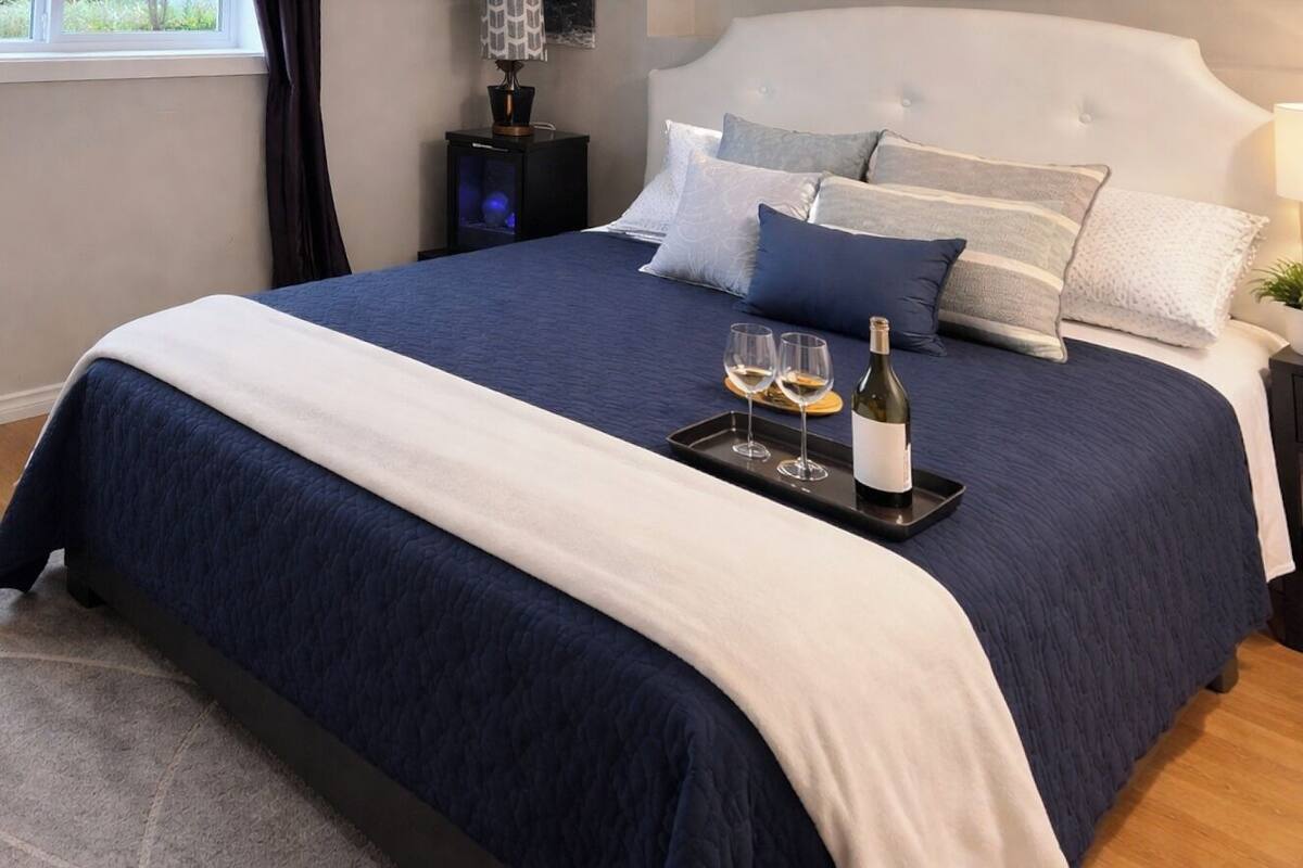 Una cama bien hecha con una colcha azul oscuro y un plaid blanco, una bandeja con dos copas de vino y una botella, y una mesita de noche con una lámpara.