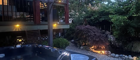 A hot tub with a water fountain, surrounded by a garden with a stone wall and a wooden pergola.