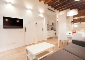 Una sala moderna con televisor de pantalla plana, un sofá blanco, una mesa de comedor y una zona de cocina con microondas y lavadora.