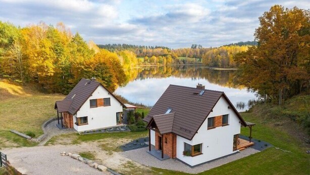 Deux maisons modernes aux toits inclinés, situées sur une pelouse, avec un lac en arrière-plan.