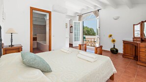 Un dormitori amb un llit, una tauleta de nit, una làmpada, un armari de fusta i vistes a un balcó amb taula i cadires.