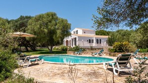 Una piscina amb gandules, un edifici blanc amb arcs i vegetació al voltant.