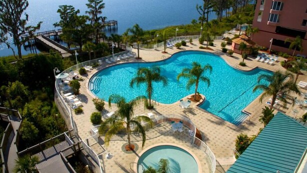 A large, curved swimming pool with a surrounding deck, palm trees, and a view of the sea. - Living the Disney Dream! | Lake View Luxury Condo (Orlando)