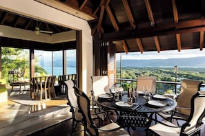 A dining area with a table set for four, large windows offering a view of the landscape, and a wooden ceiling structure.