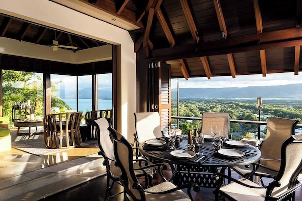A dining area with a table set for four, large windows offering a view of the landscape, and a wooden ceiling structure.