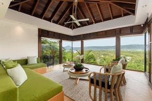 A spacious room with a green sofa, wicker chairs, a ceiling fan, and large windows offering a view of greenery and hills.