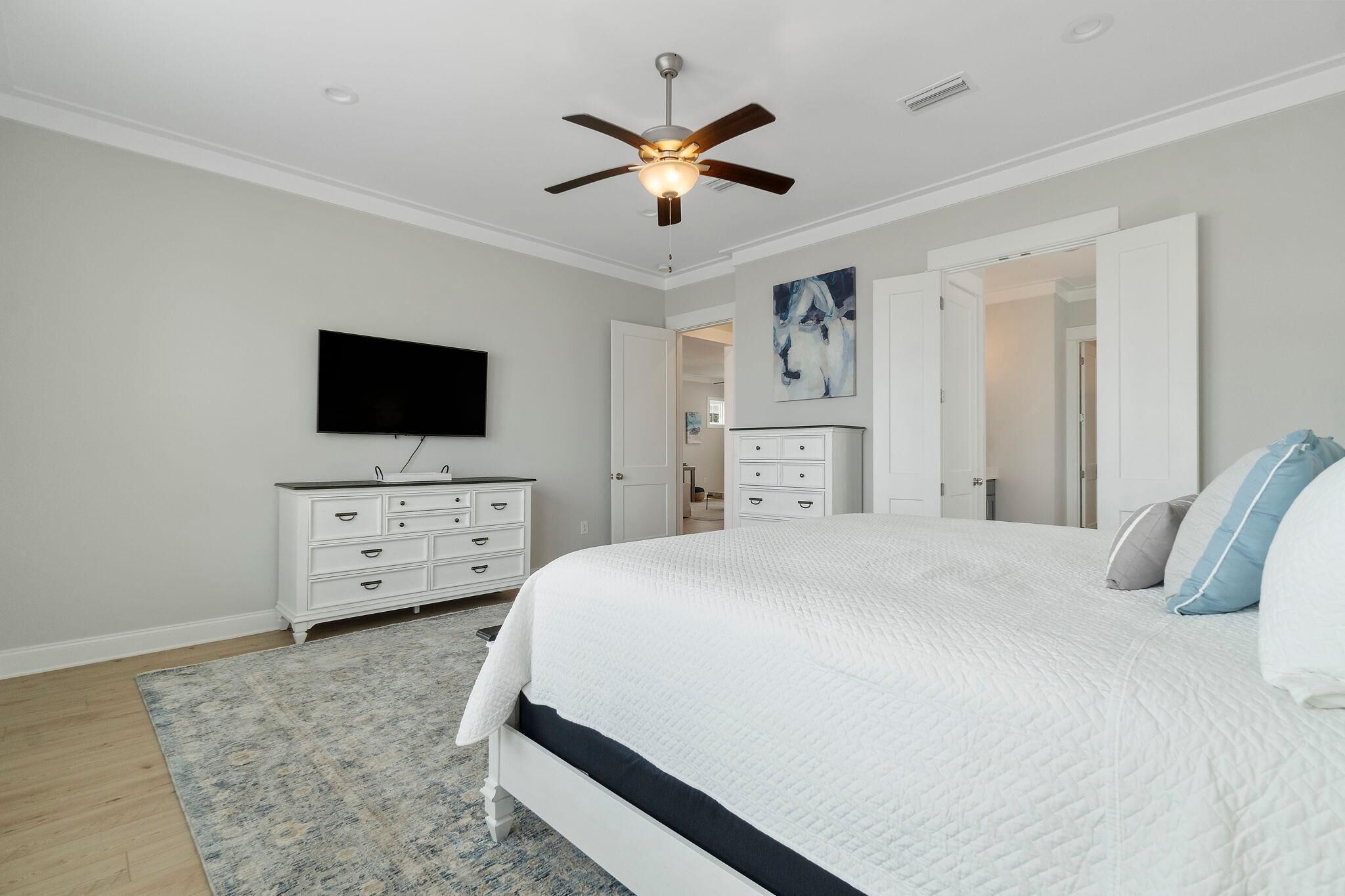 A bedroom with a bed, a ceiling fan, a flat-screen TV, a dresser, and a painting on the wall.
