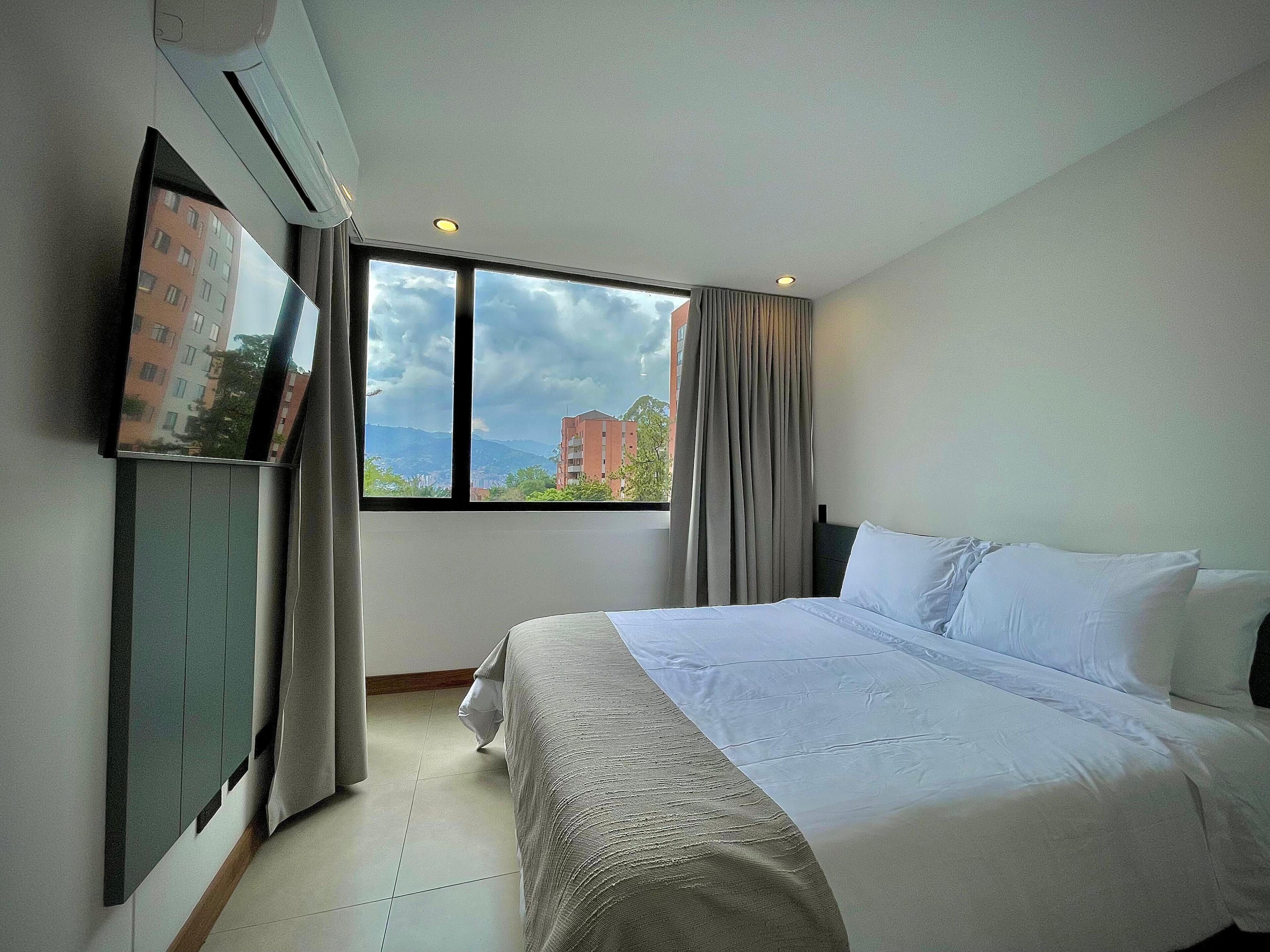 A hotel room with a large bed, a flat-screen TV mounted on the wall, and a window with a view of buildings and mountains.