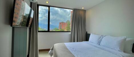 A hotel room with a large bed, a flat-screen TV mounted on the wall, and a window with a view of buildings and mountains.