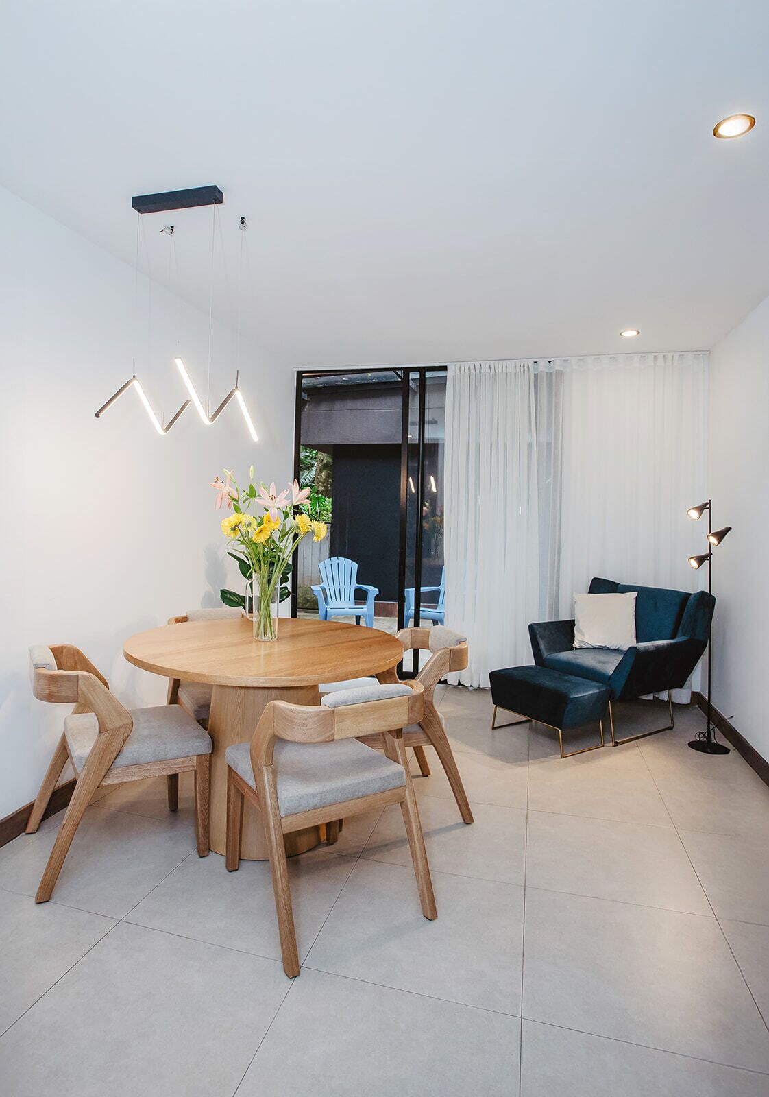 A modern living room with a round wooden dining table, light-colored chairs, a dark blue sofa, and a floor lamp.