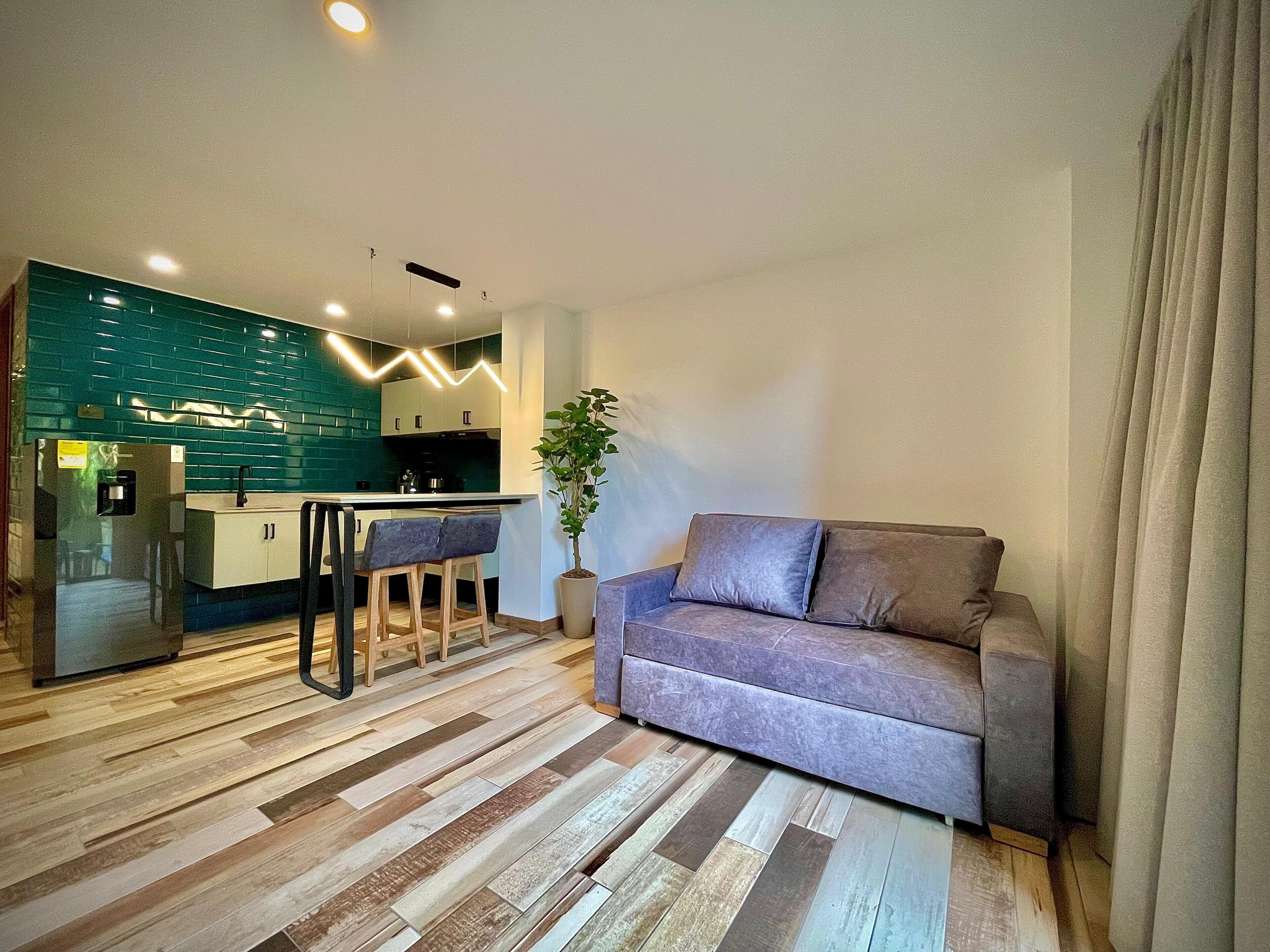 A modern living room with a grey sofa, wooden flooring, and a kitchen area with teal backsplash and white cabinets.