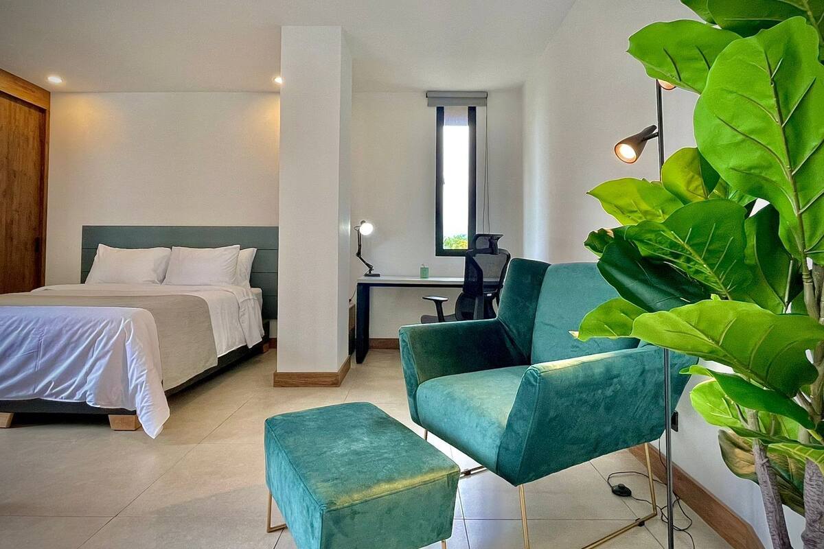 A modern hotel room with a bed, a desk, a chair, and a large green plant.