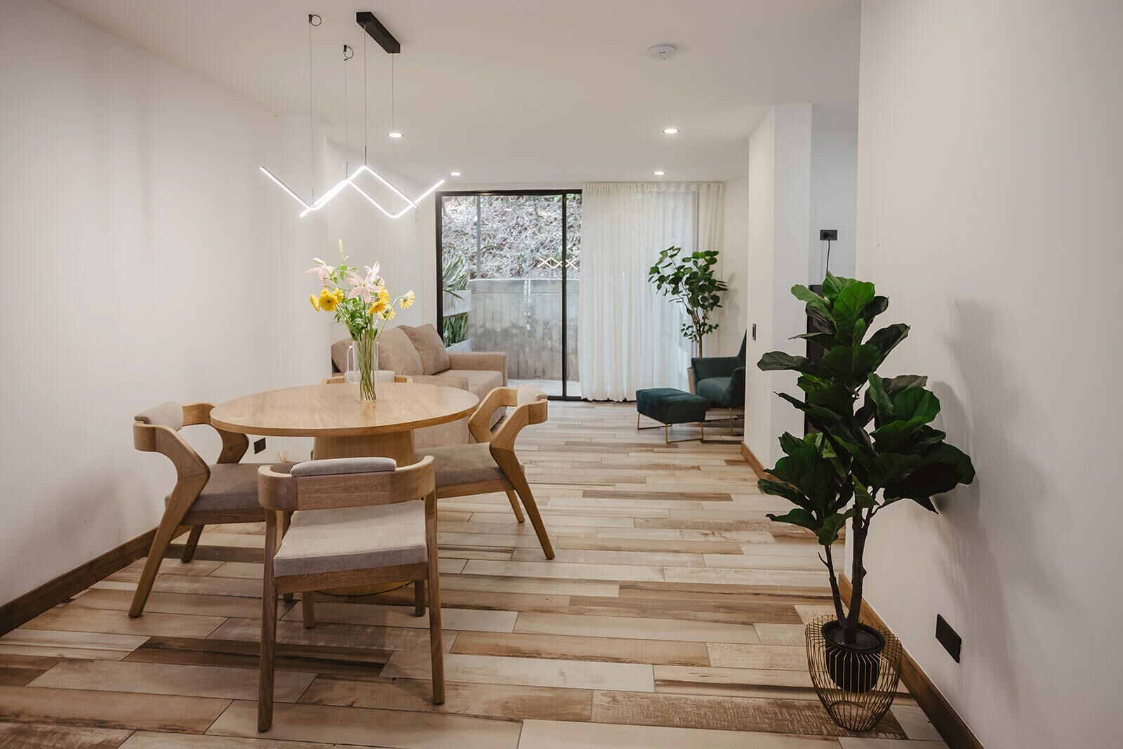 Un espace repas moderne avec une table ronde en bois et des chaises, un vase de fleurs et une grande fenêtre avec des rideaux.