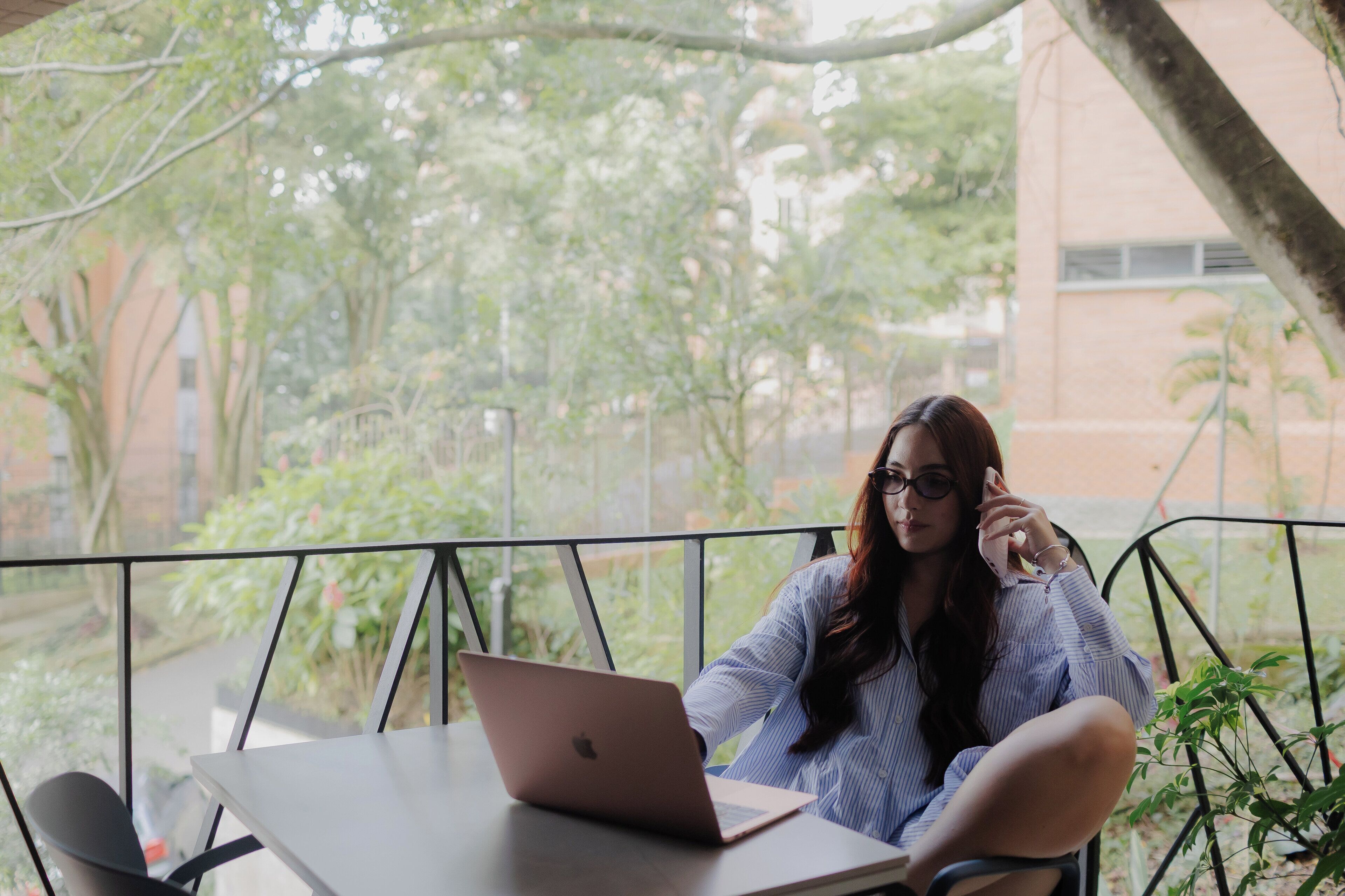 A person sitting at a table with a laptop, outdoors with greenery and a building in the background.