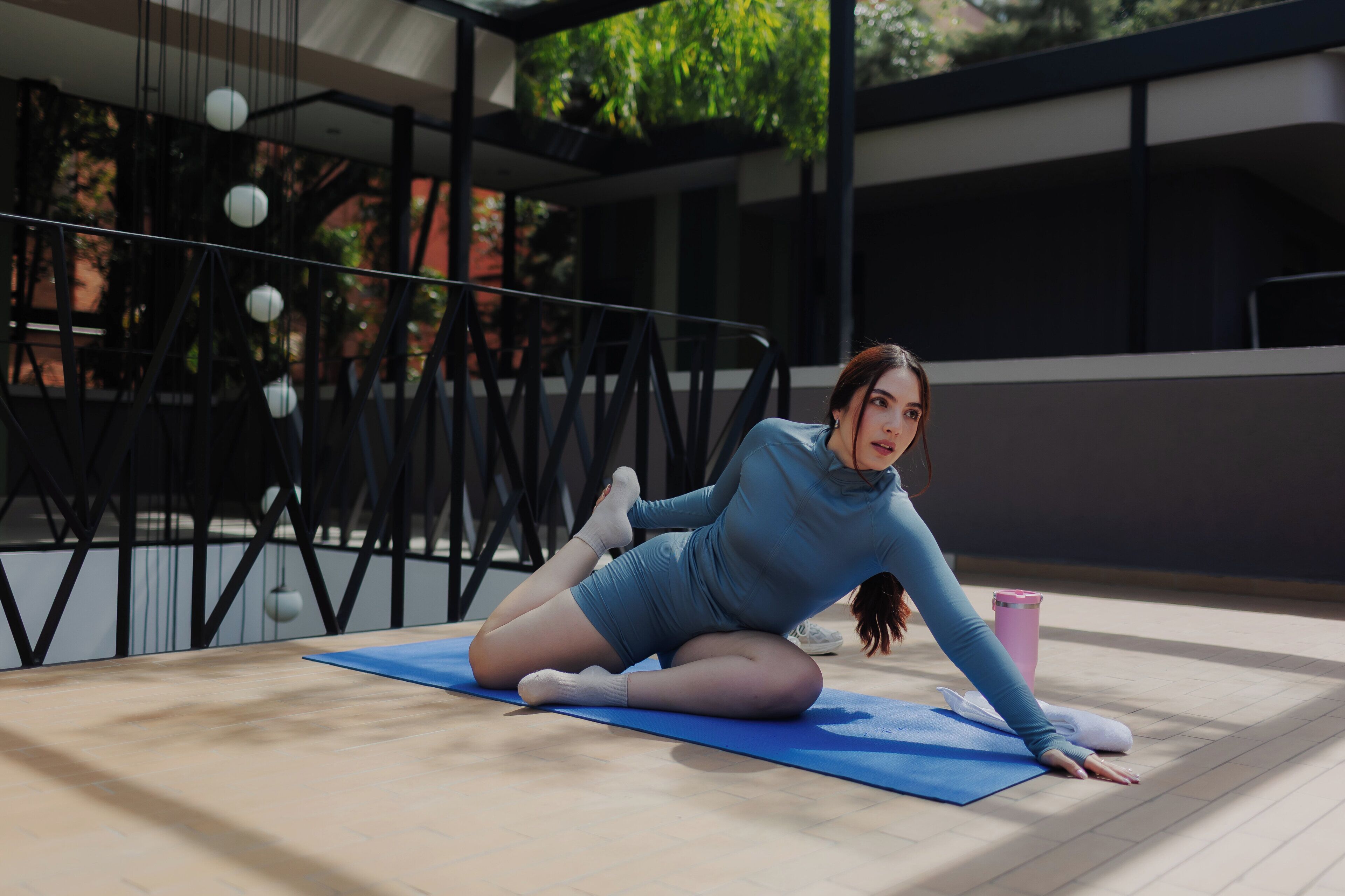 Una persona realiza una postura de yoga sobre una esterilla en un espacio exterior, con una estructura metálica y follaje de fondo.