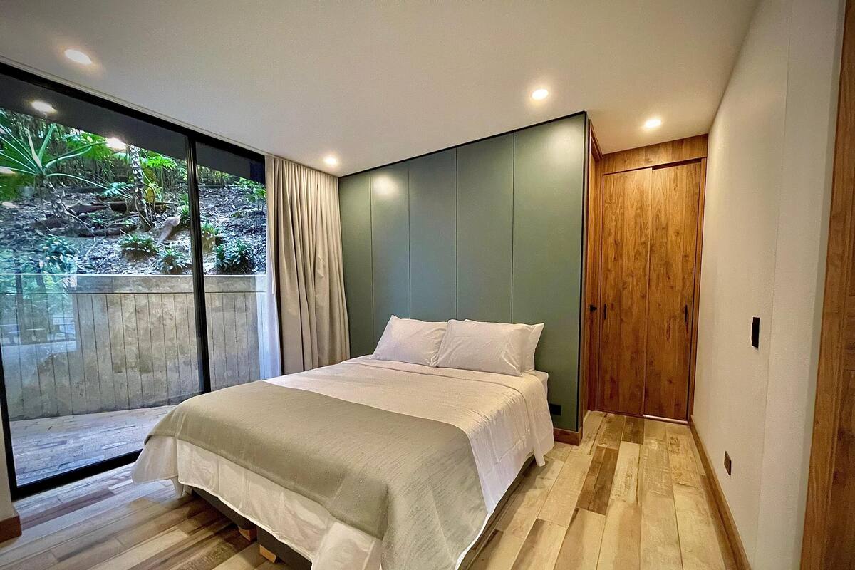 A modern bedroom with a large bed, a sliding glass door leading to a garden, and a wooden wardrobe.