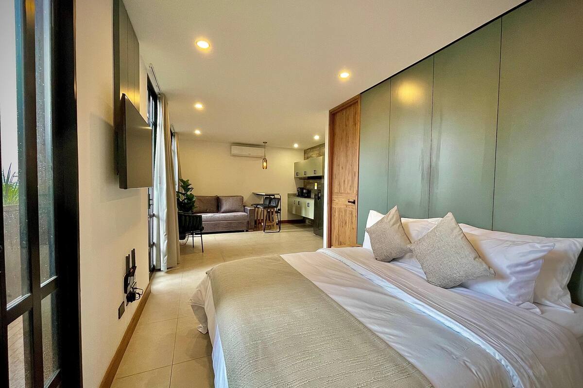 A modern hotel room with a large bed, a sofa, a dining area, and a kitchen in the background.