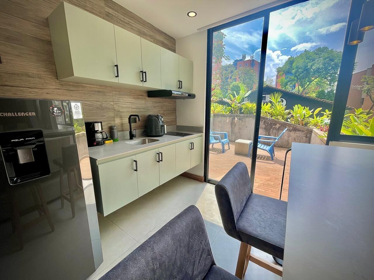 A modern kitchen with a built-in stove, coffee maker, and sink. There is a dining area with a table and chairs, and a view of a garden through large windows.