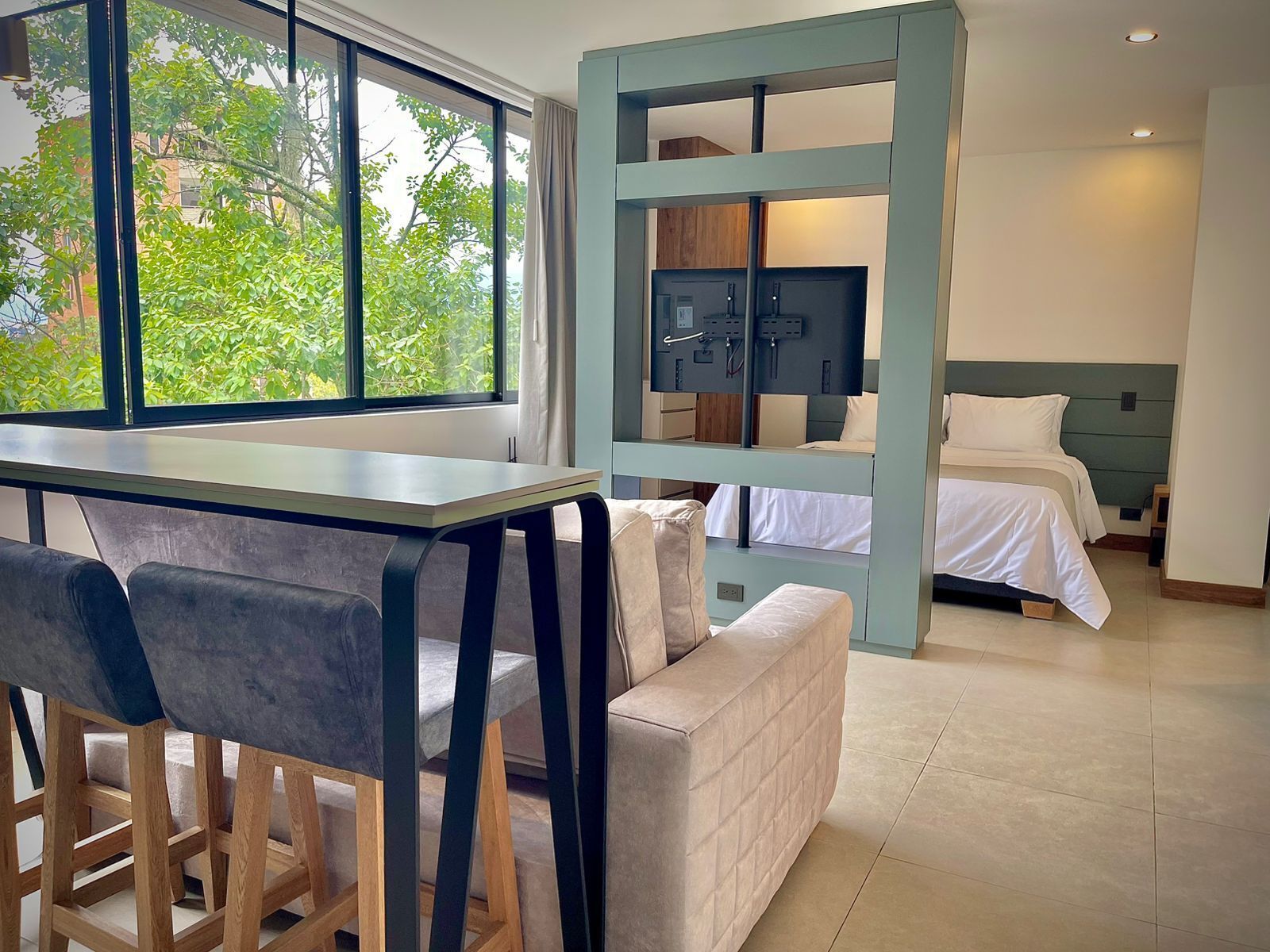 A modern hotel room with a large window, a dining area with a table and chairs, a sofa, a bed with a headboard, and a TV mounted on the wall.