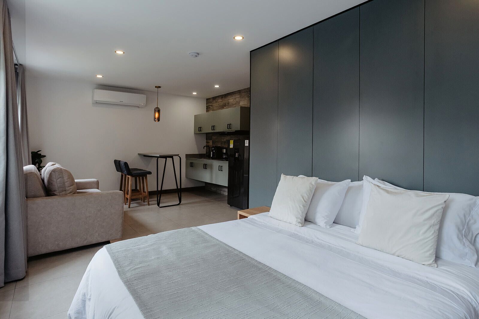 A modern hotel room with a large bed, a sofa, a dining area, and a kitchen in the background.
