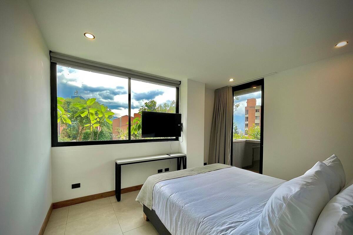 A modern hotel room with a large bed, a desk with a flat-screen TV, and a window offering a view of greenery and buildings.