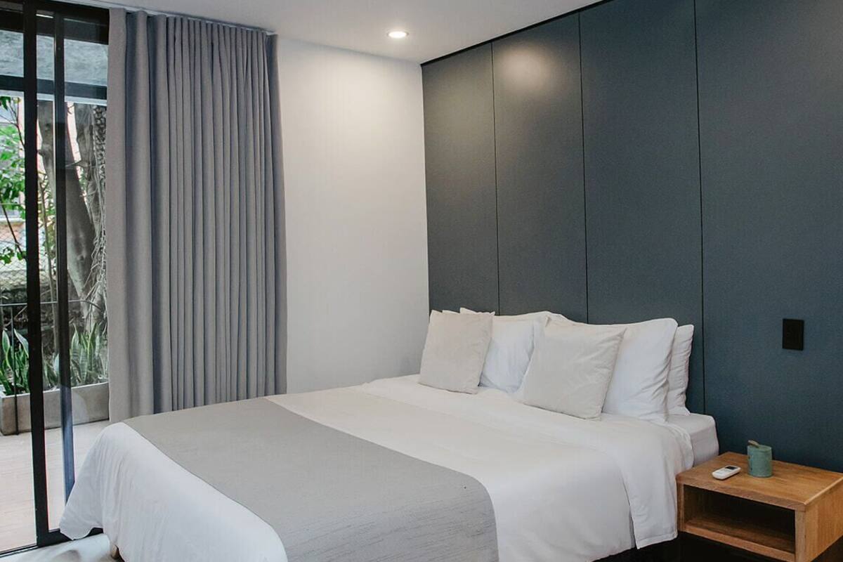 A modern bedroom with a large bed, grey curtains, and a wooden nightstand with a lamp.