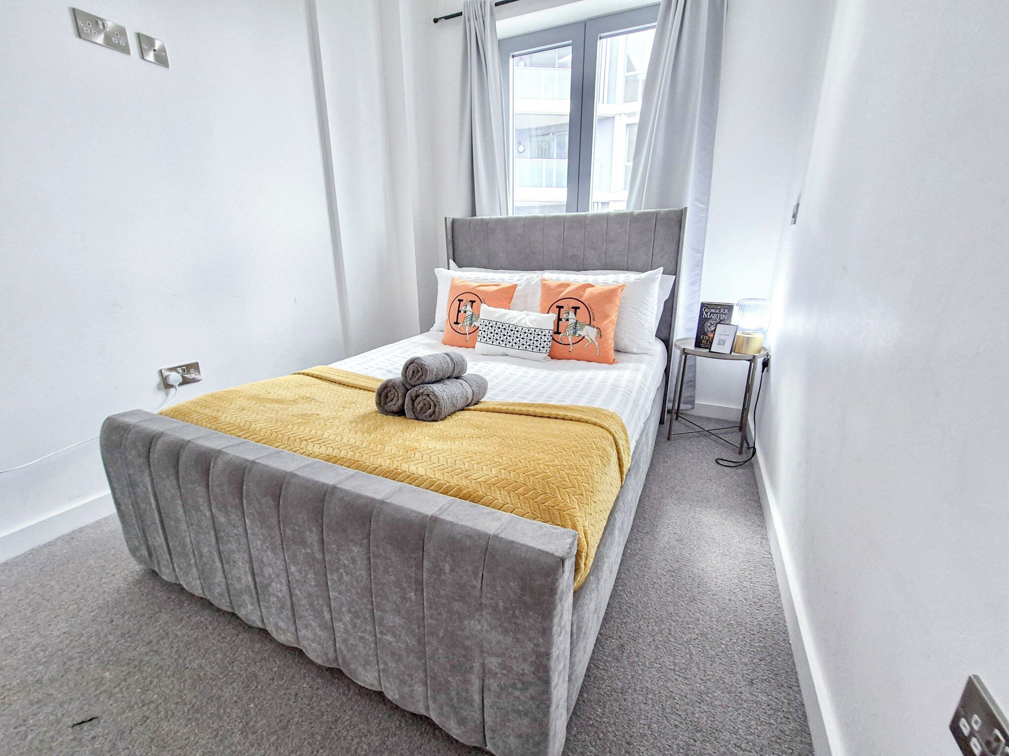 A neatly arranged bed with a grey headboard, a yellow throw blanket, and rolled towels.