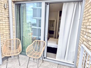 A balcony with two wicker chairs, a sliding glass door leading to a bedroom with a bed and a nightstand, and a view of the cityscape. - Lux 3-Bedroom Battersea Penthouse w/ 3 Private Balconies near Clapham, Nine Elms (London)