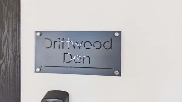 A metallic sign with the text 'Driftwood Den' mounted on a wall.