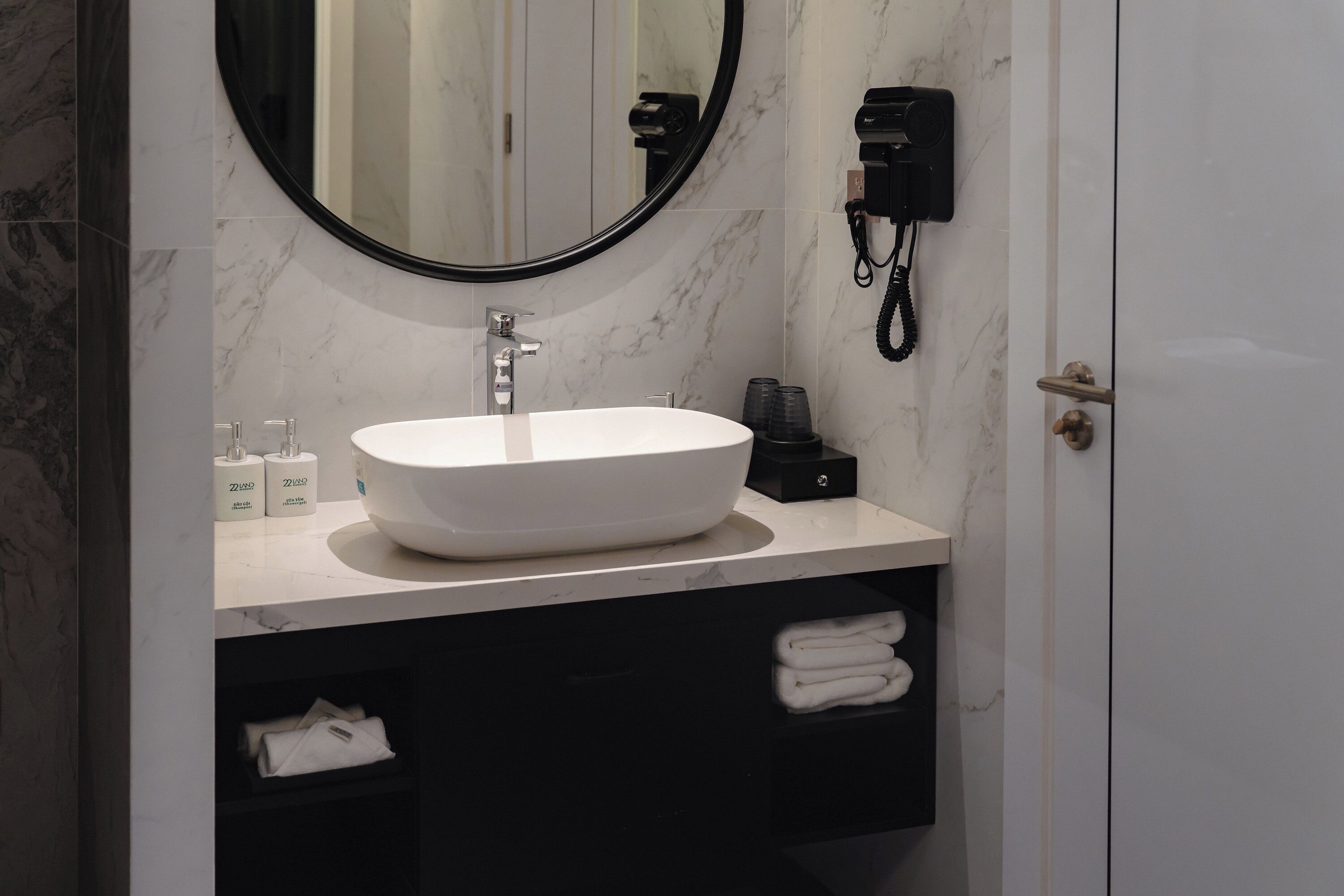 A modern bathroom with a white marble sink, a round mirror, a hairdryer, and neatly folded towels.