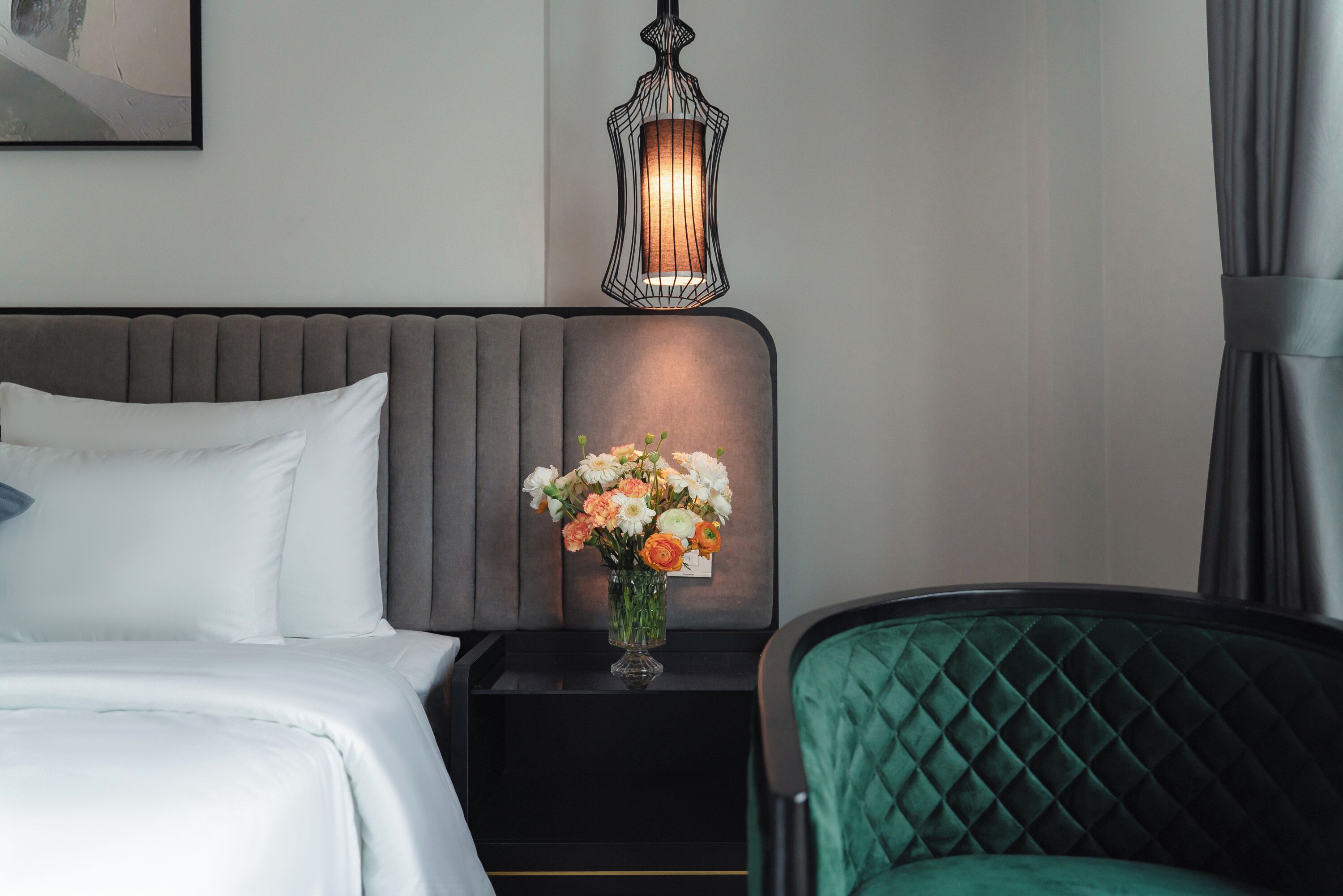Una habitación de hotel con una cama, una mesita de noche con un jarrón de flores, un sillón tapizado en verde y una lámpara de pared.