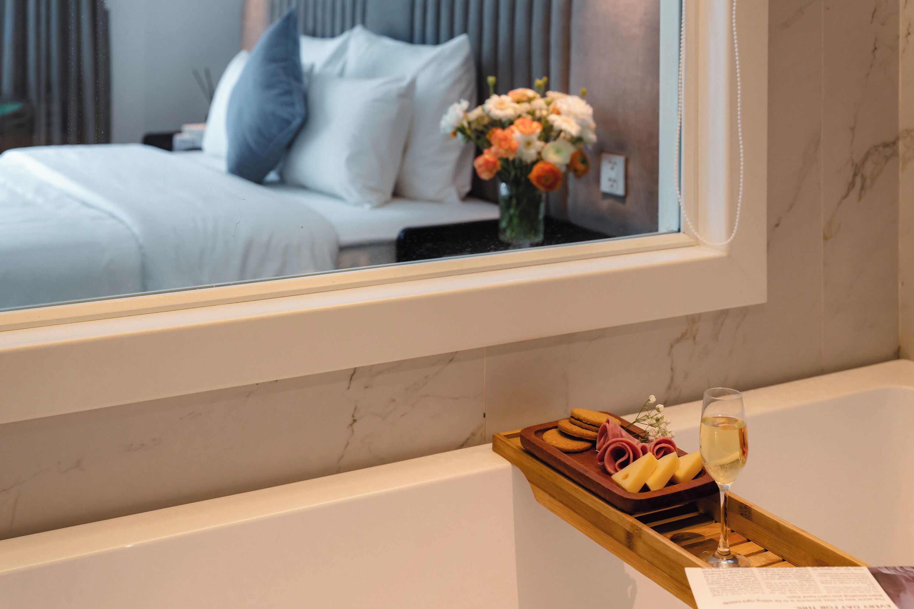 A bathroom with a bathtub, a tray with bread and a glass of wine, and a mirror reflecting a bed with pillows and flowers.