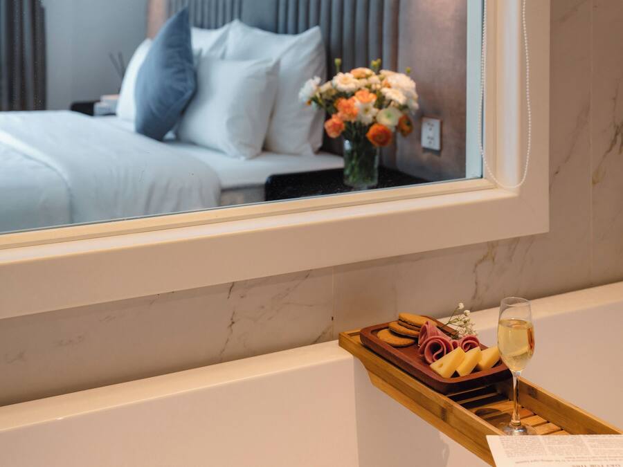 A bathroom with a bathtub, a tray with bread and a glass of wine, and a mirror reflecting a bed with pillows and flowers.