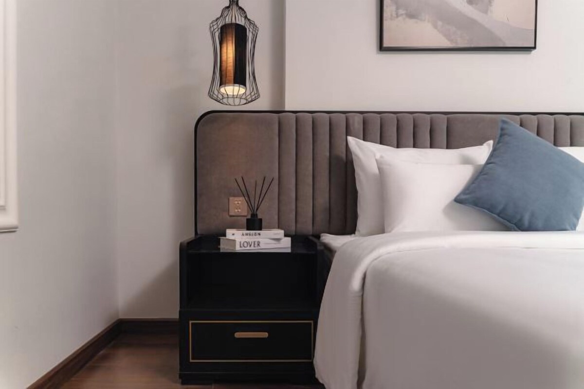 Una cama bien hecha con almohadas blancas y azules, un cabecero oscuro y una mesita de noche con una lámpara y libros.
