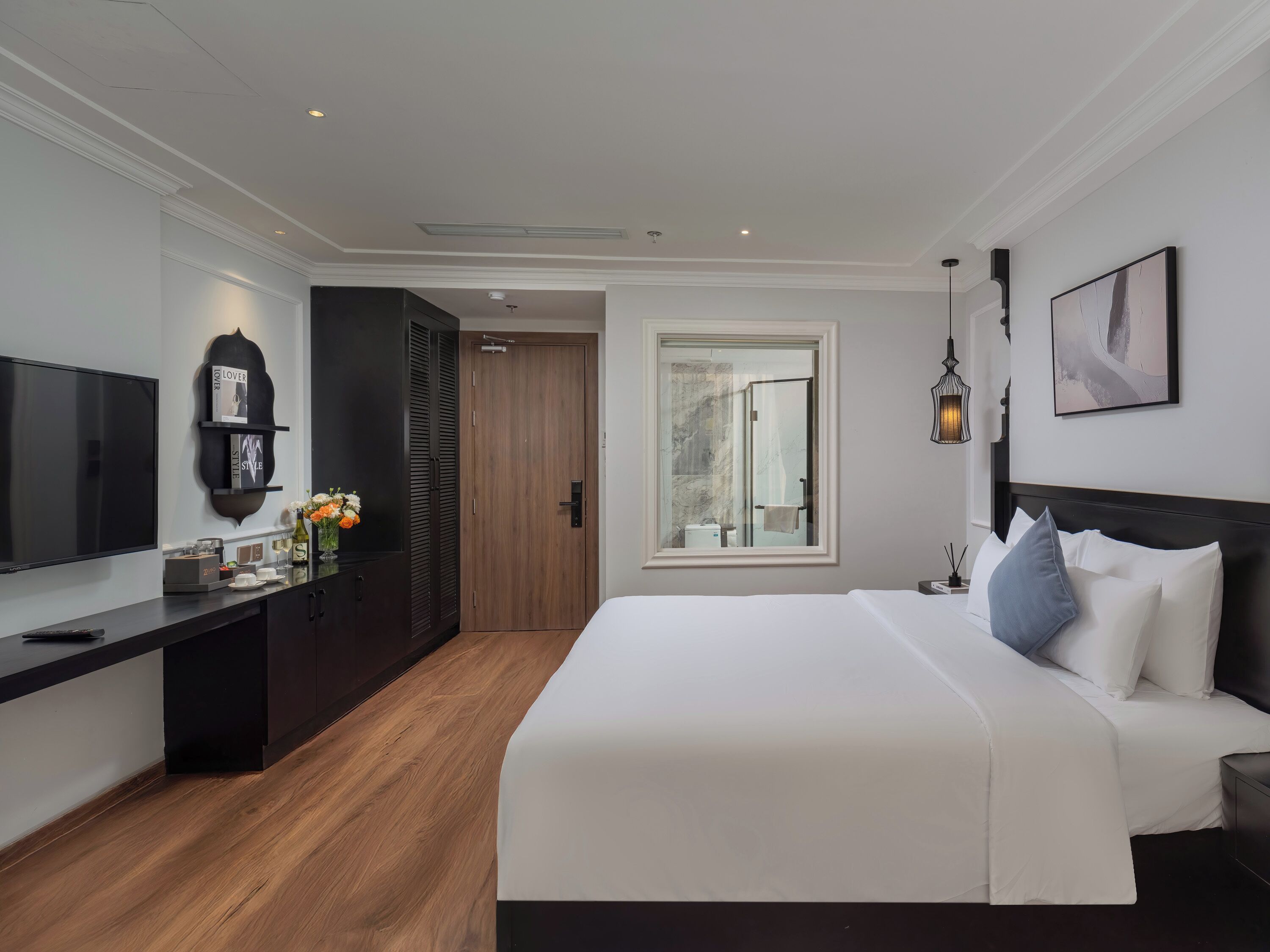 A modern hotel room with a large bed, a flat-screen TV, a dark wooden desk, and a large window.