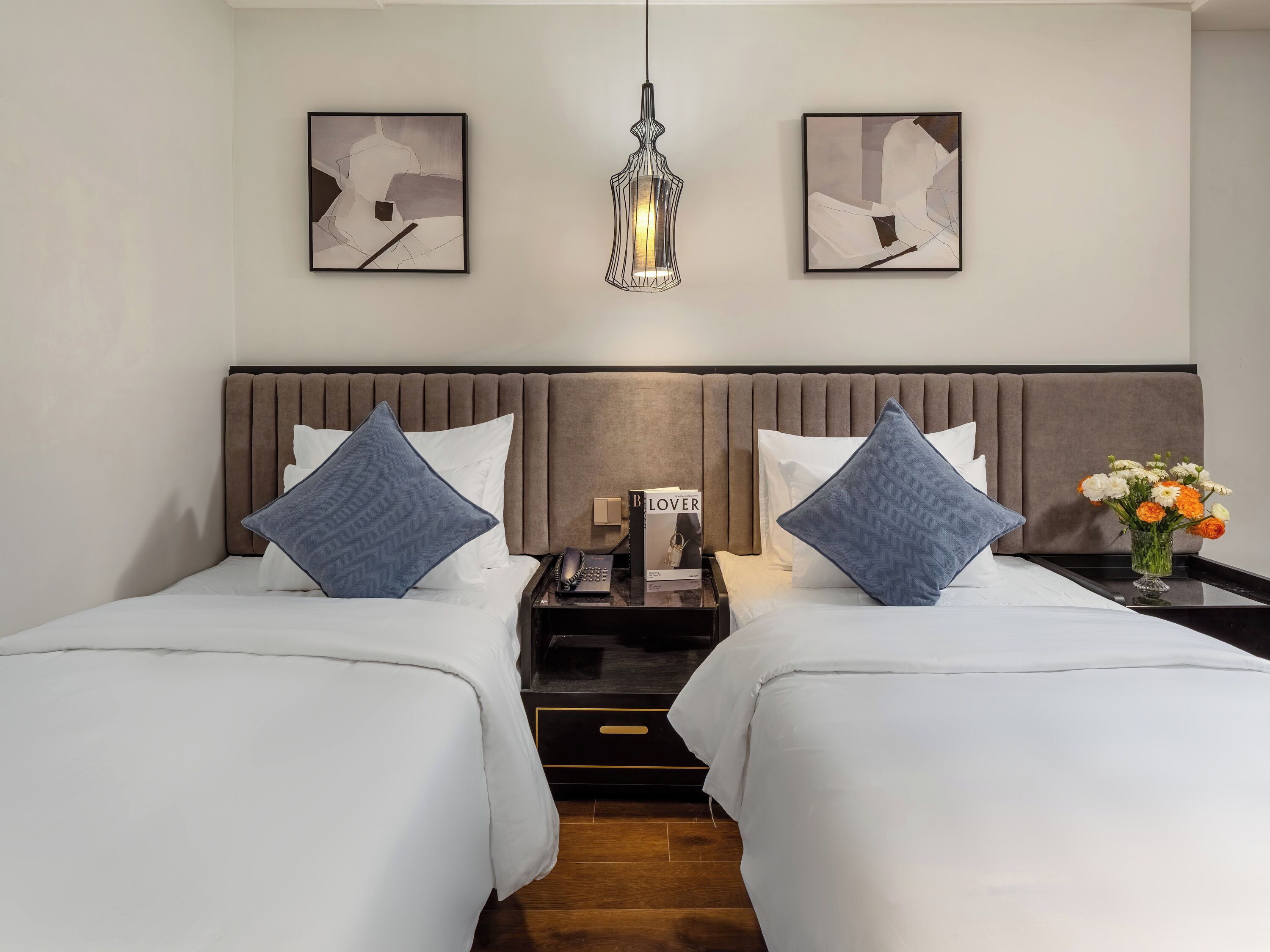 A hotel room with two beds, a nightstand, a phone, a book titled 'Lover', and a vase with flowers.