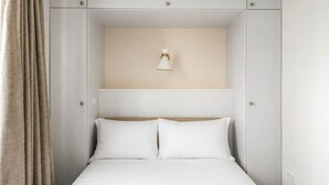 Une chambre à coucher avec un lit, deux oreillers, une table de chevet, une lampe fixée au mur et un rideau.