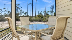 Mesa de vidrio con cuatro sillas en un espacio al aire libre techado, con vista a un paisaje arenoso con vegetación escasa y árboles altos.