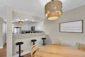 A modern kitchen with white cabinets, a granite countertop, and a stainless steel refrigerator. There is a wooden dining table with four chairs and a bar stool area with two stools. A decorative light fixture hangs above the kitchen island.