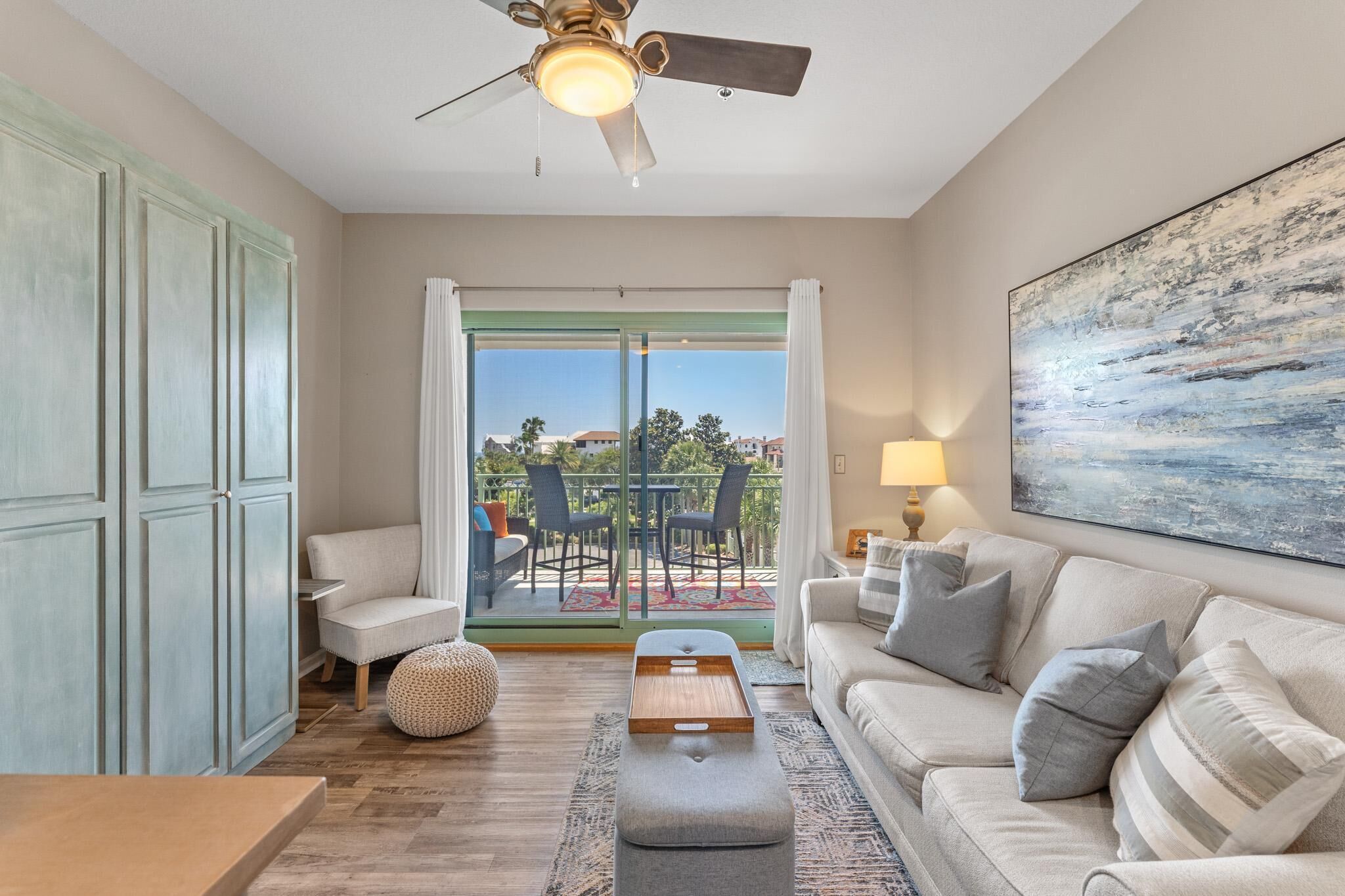 Un salon comprenant un canapé, un pouf et une table basse. On y trouve également un ventilateur de plafond, un tableau accroché au mur et, à travers la porte vitrée, une vue sur une terrasse extérieure.