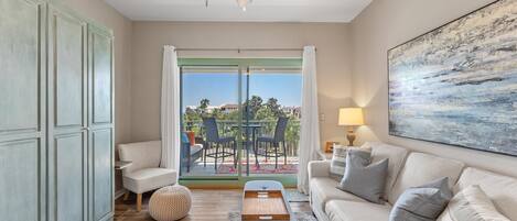 Un salon comprenant un canapé, un pouf et une table basse. On y trouve également un ventilateur de plafond, un tableau accroché au mur et, à travers la porte vitrée, une vue sur une terrasse extérieure.