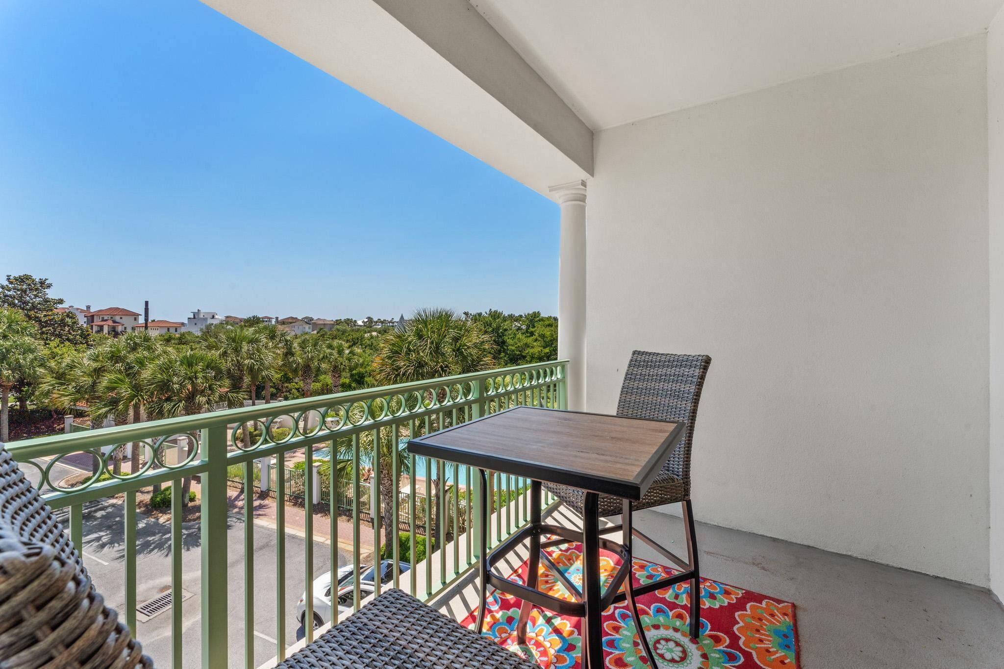 A balcony with a table and chairs, a colorful rug, and a view of a residential area with palm trees and houses.